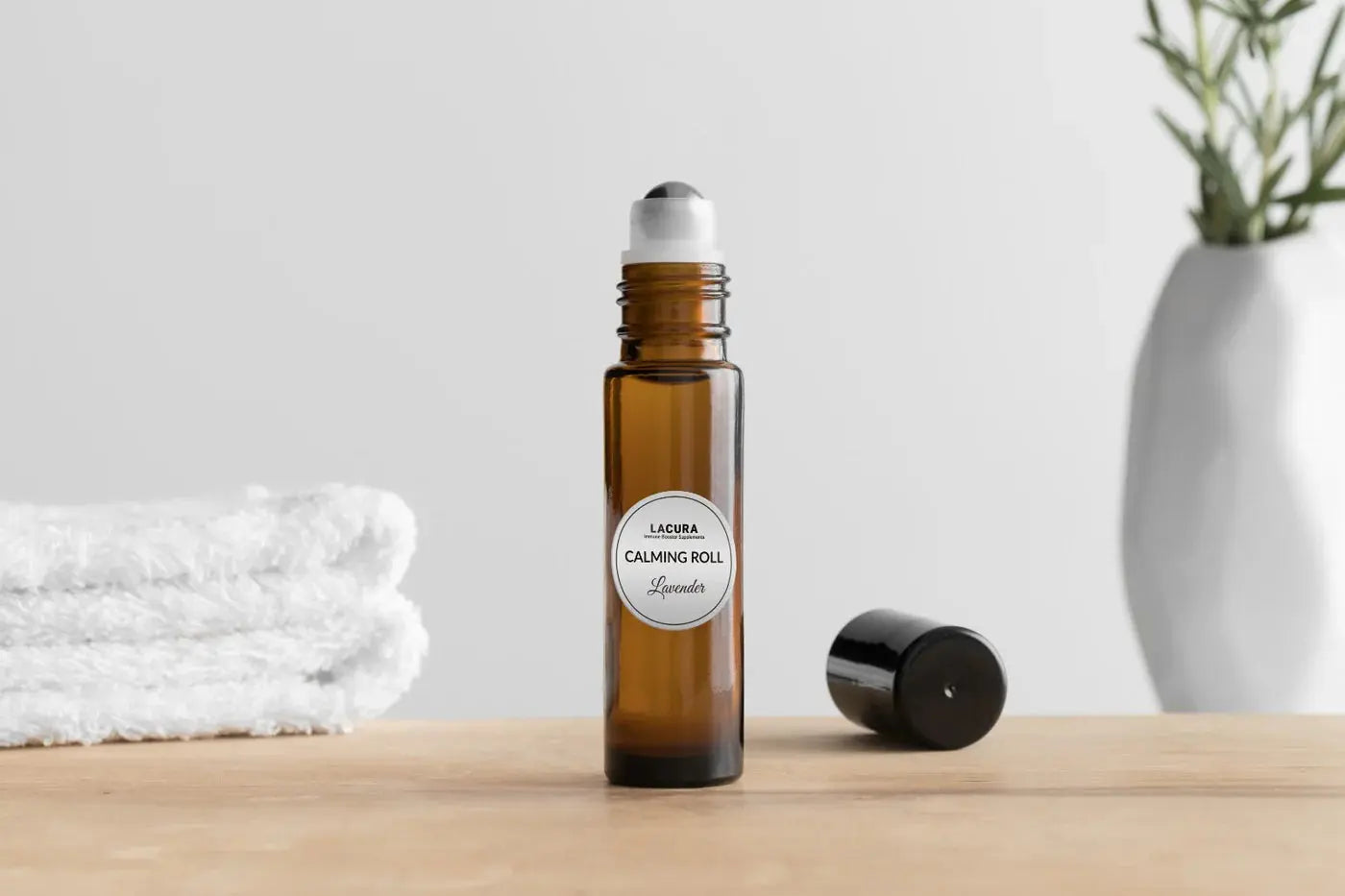 Brown roller bottle labeled 'Lavender Calming Roll' on a wooden surface with a white towel and plant in the background.
